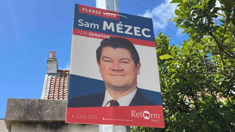 BBC An blue, white, and red election poster tied to a lampost shows a white man in a suit smiling. He has medium length black hair, slicked to one side. The poster says "Please vote Sam Mezec for Senator." It also mentions his party, Reform Jersey, and the election date - June 7th 2026.