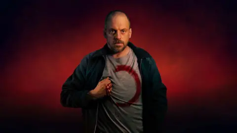 Actor Matthew Gurney wearing a T-shirt with a red unfinished circle and his bloodied hand on his chest, against red background.