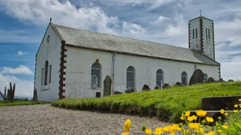 MANX SCENES Jurby Church is a white painted building with a tower on the right-hand side. There is a gravel path around it and yellow poppies on the right of a set of steps leading up to the green grass of the churchyard.