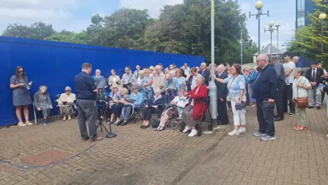Church of Scotland An open air church service, where a minister speaks to a crowd of parishioners standing outside, with a large blue fence blocking off entry to the damaged church that is nearby.