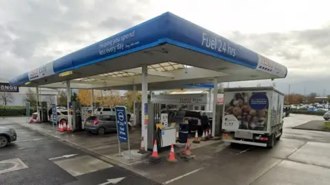 A petrol station with a blue roof and a number of vehicles at the pumps including a small lorry