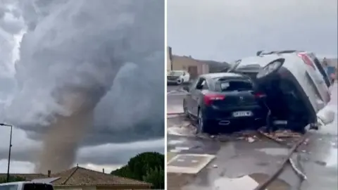 A tornado is seen spinning above a house in the clouds, with two cars on top of each other on the right.
