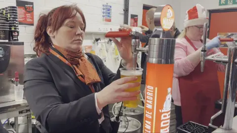 A woman in a dark jacket serving beer from a tap. Another woman in an Aberdeen supporters hat is serving beer in the background