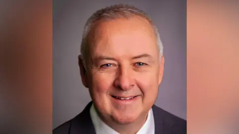 A man with short white grey hair is smiling. He is wearing a white shirt and black suit jacket