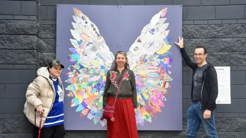Three people stand around the artwork made of feathers that are placed in a way that makes them look like a pair of wings. One woman stands in the middle, as if they are her wings, and is smiling at the camera. A man and woman stand on either side of the piece, also smiling at the camera.
