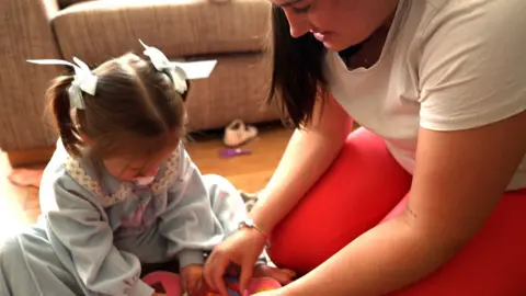 Florence Woman kneeling on the floor playing with a little girl with hair up in bunches wearing a long sleeved light blue dress. The woman wears red leggings and a white t-shirt and has dark brown long hair.