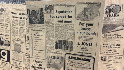 Old, yellowing newspapers that include articles about the carpet shop being open for 50 years. One of the headlines says "Reputation has spread far and near!". There are black and white photographs of people inside the carpet shop.