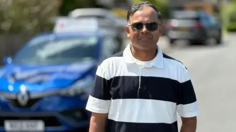 Deen poses in front of his car. He has short black/grey hair and wears sunglasses and a black and white short sleeved collared shirt.