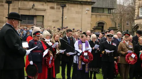 Lancaster City Council About a dozen dignitaries including a lady mayoress and military leaders hold poppy wreathes and sing hymns at the service with crowds and soldiers in camouflage uniform in the backdrop.