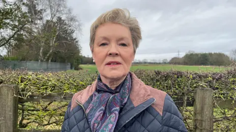 Susan pictured looking into the camera with a neutral expression. She has short blonde hair, and is wearing a navy blue quilted jacket with light brown suede patches on the collar and shoulders. She's also wearing a dark blue scarf with a purple, red and green paisley pattern on it. Susan is stood in front of a hedge, which is bracketed by a wooden fence. Behind her, a grassy field stretches into the background, with a row of trees running along the left side of the photo. In the far distance, and slightly out of focus, electricity pylons can be seen. The sky is grey and overcast. 