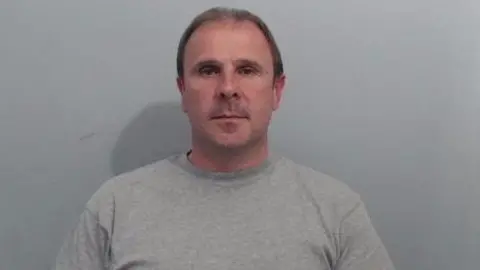 PSNI A mug shot of Irvine. It is a head and shoulders image, he is wearing a grey sweatshirt and has short brown hair. He looks directly at the camera. Behind him is a plain grey wall.