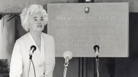 The Open University Jennie Lee standing at a podium setup with three microphones arranged on stands in front. She is wearing a light‑coloured, formal jacket with distinctive knot-style fastenings on the front. Behind is a large commemorative plaque mounted on a wall. The plaque includes engraved text and an emblem at the top, indicating it marks the opening or dedication of a facility or building. 