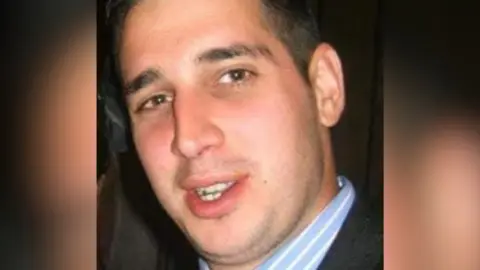 Martin smiles in a close-up photo. He has dark brown hair and dark eyebrows, and wears a light blue shirt with white stripes and a dark suit jacket. 