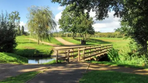 Worcestershire County Council A wooden bridge over a waterway, it is sited in a park with trees behind it and a pathway on either side