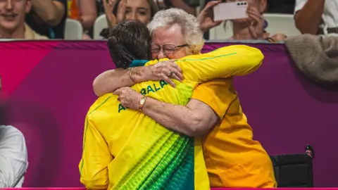 An athlete in a yellow and green tracksuit is hugged by a woman with grey hair, wearing an orange shirt. The other spectators are warmly cheering and clapping.