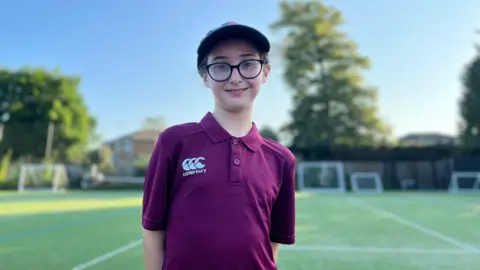 Rosina is wearing a purple shirt and a baseball cap and is behind a pitch.