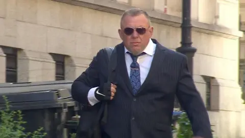Mark Killick wearing a white collared shirt, a blue paisley tie and a dark navy pinstripe suit. He is walking into Bristol Crown Court with brown sunglasses on and a black rucksack slung over his right shoulder. 