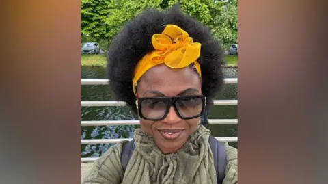 OLUWASANYA FAMILY  Dark haired woman with yellow hair-tie. Wearing black sunglasses and a khaki green jumper. 