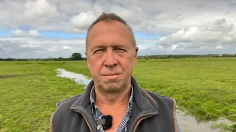 Mike Curtis, a middle-aged man with short brown hair standing in a muddy green field. He's wearing a fleece gilet with a checked shirt and is looking at the camera.