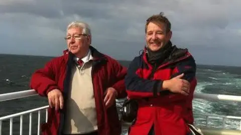 Two men - Stuart Flett and his dad Rodney - standing on a ferry, with the sea behind them.