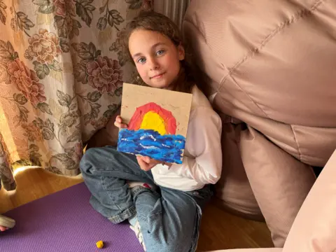 A young girl holds up a piece of art she has produced , it is a rainbow over the sea