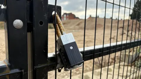 Martin Heath/BBC A black steel gate, with a padlock across the opening. There is a landscape of what appears to be sand behind the gate.
