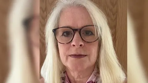 Close up of Sue's face, she has blonde hair and black glasses with brown eyes. She is stood in front of a wooden frame.
