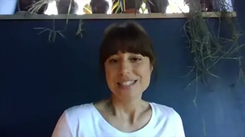 Zahra Ahmadi looking at the camera and smiling. She is sat against a blue wall with a hanging plant on the right.