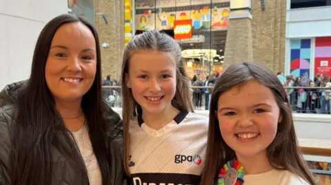 Aoife, Niamh and Saoirse standing side by side. Aoife has long dark hair and is wearing a green coat. Niamh has long brown hair which is partially tied back and is wearing a sports top. Saoirse has long brown hair and is wearing a white top.