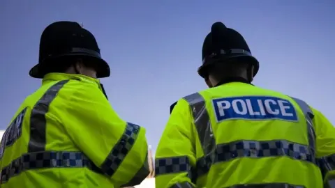 Two police officers stand with their backs facing the camera. They are wearing black police hats and yellow hi-vis police jackets.