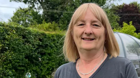 Helen has light-ginger hair and wears a blue T-shirt and silver necklace. She is standing in a garden in front of green hedge.