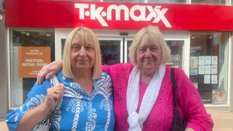 BBC A woman with blonde hair is on the right wearing a pink top and has her right arm around another woman with blonde hair on the left, who is wearing a blue and white top. They are in front of TK Maxx.