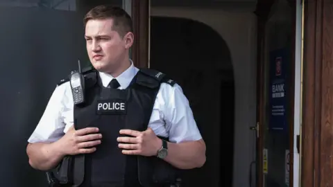 Finnian Garbutt, a man with dark hair, wearing a white shirt with black tie and a black support vest with the words police on it. He is wearing a black watch on his left wrist and there is a brown wooden door in the background.
