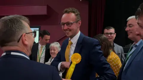 Image of Adam Dance. He has light hair, facial hair and glasses. He is wearing a suit, yellow tie and a yellow Liberal Democrat badge. 
