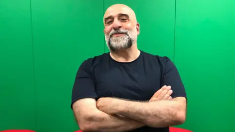 Comedian Omid Djalili is wearing a black T shirt and has his arms folded. He stands in front of a grey background with a slight smile 