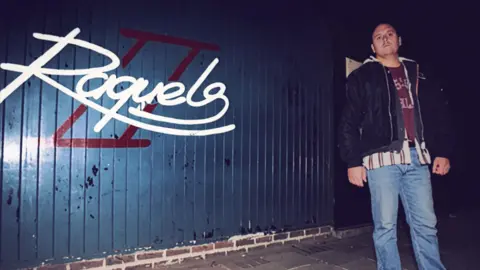 Bernard O'Mahoney Bernard O'Mahoney wearing jeans, a shirt and a black jacket outside Raquels. The club's name is printed in white font on a blue wall behind him.