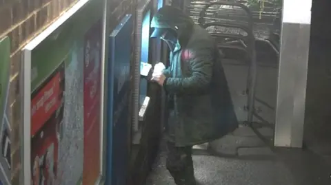 ERSOU A person wearing a hooded coat and cap is using an outdoor ATM at night. The scene is dimly lit, with bright light coming from the ATM screen. There are posters on the brick wall to the left and a stack of shopping carts in the background.