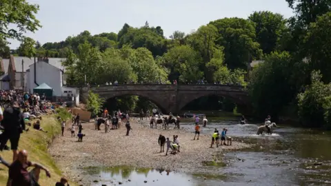 PA Media View downriver of people attending the Appleby Horse Fair. People are lining the banks of a river to watch riders taking their animals into the river. Others are watching from a bridge. The weather is sunny.