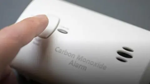 A finger pressing a button on a white carbon monoxide alarm. 