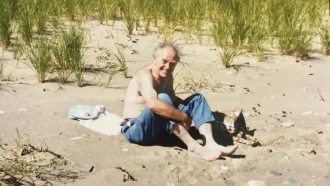 Family handout Paul, who is shirtless and is wearing jeans on a summer's day, is sitting on the sand on a beach. 