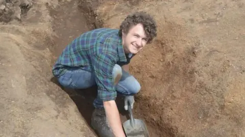 Jowan Albon Jowan Albon smiling at the camera in a muddy hole. He has a spade in his right hand and is wearing a green and blue checked shirt. 