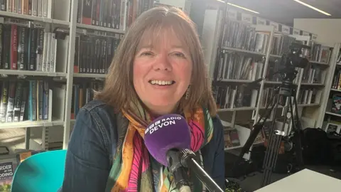 A woman sitting in a library, talking into a purple microphone. A bookshelf is behind her. She is smiling at the camera and is wearing a colourful scarf.