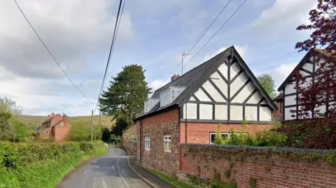 A cottage stands near a road with a walled garden. The walls are partly brick and partly black and white. Across the road, there is a grassy verge and hedge. Further cottages are around the property. There is open countryside further away.
