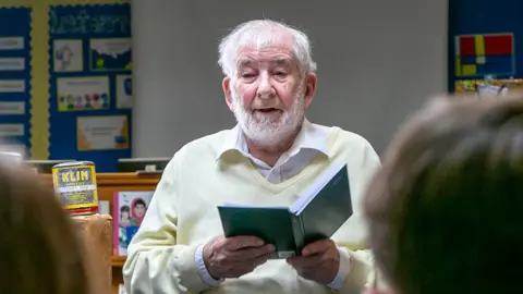 Rod Bryans Francois sits reading from a diary. Next to him on a shelf is a tin of powdered milk, with the brand name KLIM