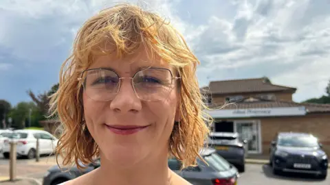 Imogen Lee/BBC Jenny Knight is standing in front of a car park. She has short ginger hair and is wearing clear framed glasses. In the background is a building which has an automatic door, and there are several cars parked outside of the building.