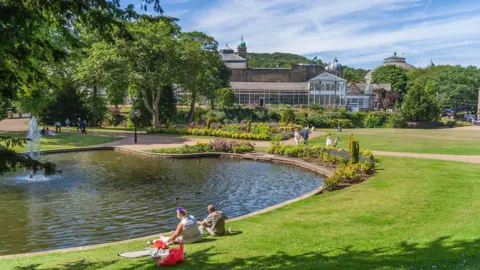 A couple sits on a lawn next to lake in the middle of a large park on a sunny day. Behind them is a large pavilion, orangery and dome-roofed building
