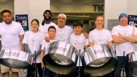 BBC Eight people from the steel band. they are in white t-shirts that say "impact steel band". They are standing in front of three silver steel drums.
