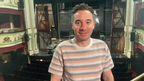 BBC/Piers Hopkirk James Graham, wearing a striped t-shirt, sits in the stalls of the Theatre Royal, Brighton, with his back to the stage.