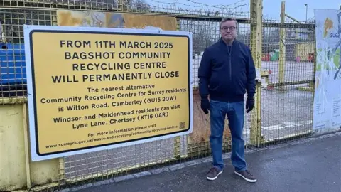Clare Cowan/BBC A man - councillor Richard Wilson - wearing a black jacket and black gloves stands to the right of a big yellow sign. The yellow sign reads: "From 11th March 2025 Bagshot Community Recycling Centre will permanently close / The nearest alternative Community Recycling Centre for Surrey residents is Wilton Road, Camberley (GU15 2QW) / Windsor and Maidenhead residents can visit Lyne Lane, Chertsey (KT16 0AR)"