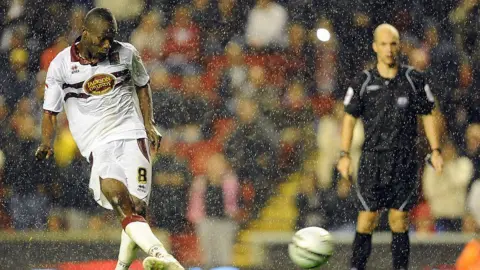 Getty Images A man in a white football kit kicks a ball in the pouring rain. A man in a black referee kit looks on. 